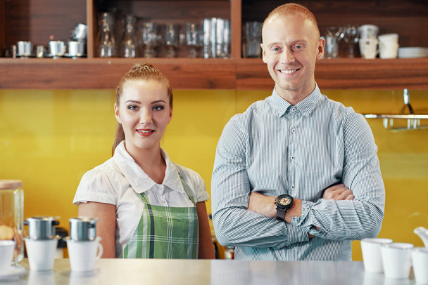 Улыбающаяся женщина в фартуке и элегантный мужчина-работодатель, стоящий вместе за стойкой в кофейне и улыбающийся в камеру
 - Фото, изображение