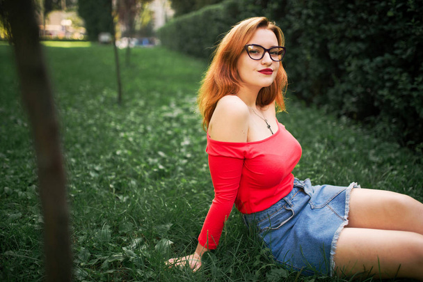 Attractive redhaired woman in eyeglasses, wear on red blouse and jeans skirt posing at green park. - Photo, image