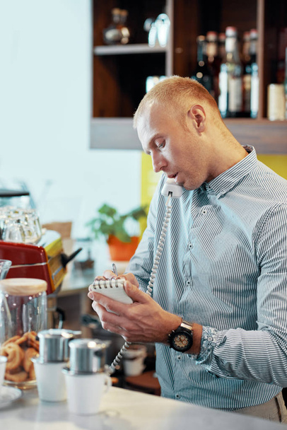 Hombre hablando por teléfono y tomando notas de pedido mientras tiene una llamada telefónica en el mostrador en la cafetería
 - Foto, imagen
