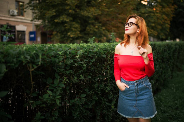 Attractive redhaired woman in eyeglasses, wear on red blouse and jeans skirt posing at green park. - Foto, Imagen