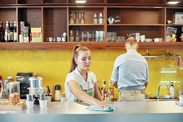 Woman working as barista and wiping counter of bar with manager standing near  - Photo, Image