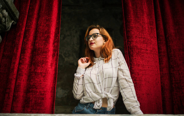 Attractive redhaired woman in eyeglasses, wear on white blouse posing at arch of open red curtains. - Fotoğraf, Görsel