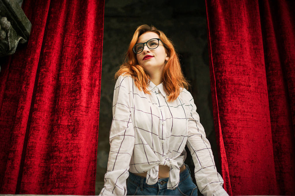 Attractive redhaired woman in eyeglasses, wear on white blouse posing at arch of open red curtains. - Foto, imagen