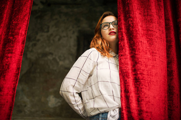 Attractive redhaired woman in eyeglasses, wear on white blouse posing at arch of open red curtains. - 写真・画像