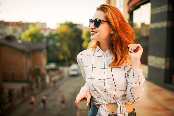 Attractive redhaired woman in sunglasses, wear on white blouse posing at street against modern building. - Φωτογραφία, εικόνα
