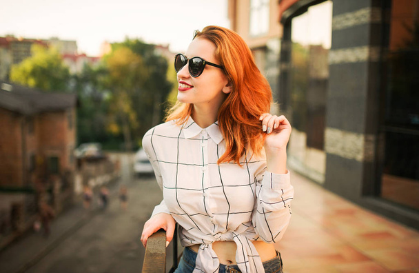 Attractive redhaired woman in sunglasses, wear on white blouse posing at street against modern building. - Фото, зображення