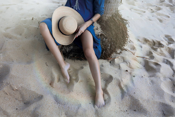 Picture of slim tanned woman's legs. Lady sitting on the palm roots on the beach, holding straw hat with black ribbon. Blue light summer dress. Warm sand and cold ocean water. Relaxing on the island. - Foto, afbeelding