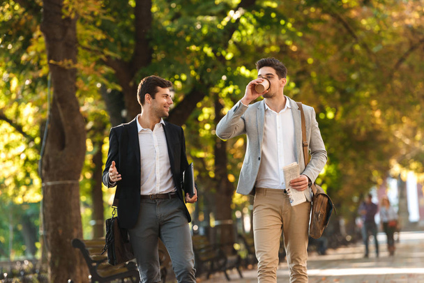 Two happy businessmen walking outdoors, drinking coffee, talking - Foto, immagini