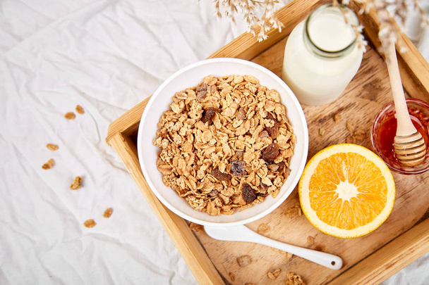 Good morning. Breakfast on white bed sheets. Muesli or granola, milk, orange  on wooden tray from above. Top view.  Flat lay. Copy space. Hotel Room Early Morning. - Photo, Image