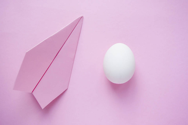 Ένα λευκό μόνο αυγό σε ροζ φόντο με αεροπλάνο χαρτί και copyspace. Έννοια του ταξιδιού. - Φωτογραφία, εικόνα
