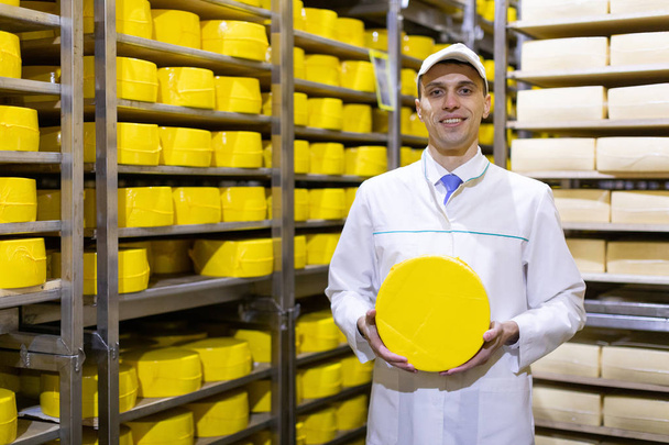 homem em um manto branco com uma cabeça de queijo em suas mãos está em um armazém de queijo
 - Foto, Imagem