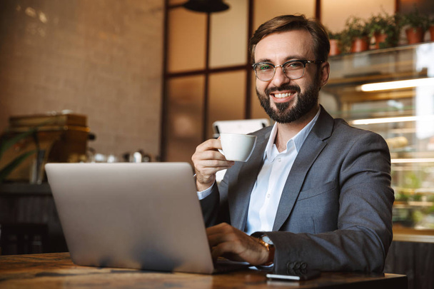 ハンサムな青年実業家を着たスーツのラップトップ コンピューターで作業して、屋内では、カフェに座ってコーヒーを飲みながら - 写真・画像