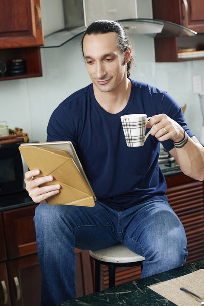 Mosolygó fiatalember iszik kávét, és olvassa híreket a tabletta számítógépen - Fotó, kép