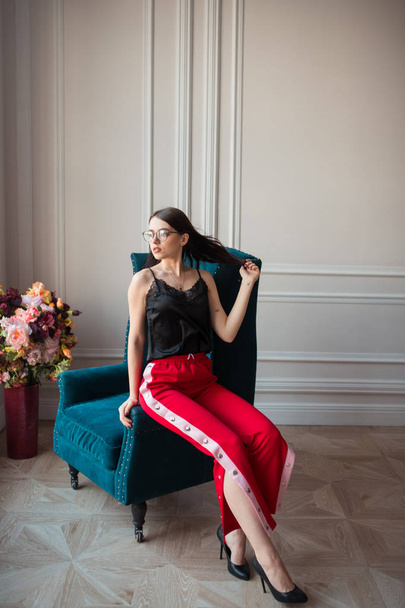 stijlvolle jongedame in brillen in rode broek poseren op fauteuil binnenshuis - Foto, afbeelding