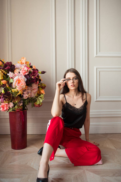 stylish fashion model in eyeglasses and red pants posing with bouquet of flowers on floor indoors - Fotó, kép