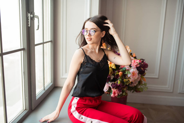 şık genç kadın kırmızı çiçekli kapalı pencere pervazına poz pantolon - Fotoğraf, Görsel