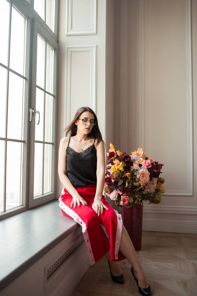 stylish young woman in red pants posing on window sill with flowers indoors - Фото, зображення