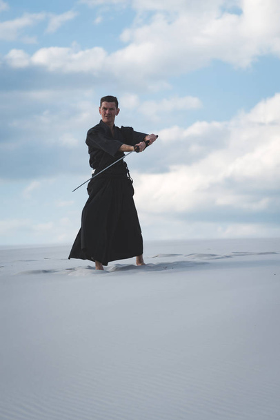 Man in traditional Japanese clothes with sword katana is training martial arts in desert during sunset - samurai on the cloudy sky background. - Foto, imagen
