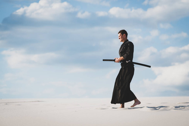 Man in traditional Japanese clothes with sword katana is training martial arts in desert during sunset - samurai on the cloudy sky background. - Fotoğraf, Görsel