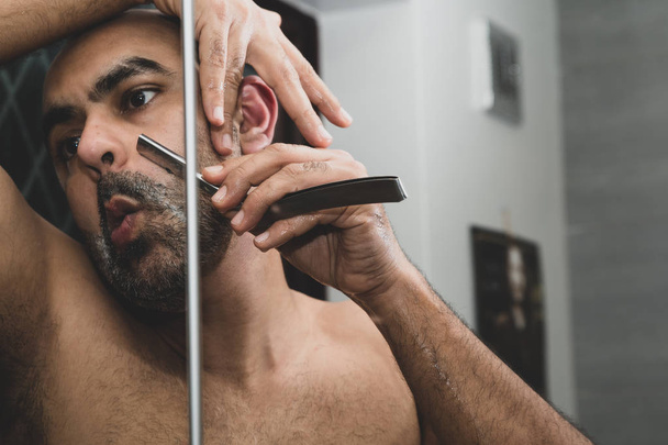 Um homem asiático britânico careca, arrumando e esculpir sua barba em casa no banheiro, em frente ao espelho. Ele está usando uma navalha de garganta cortada tradicional ou uma navalha de barbeiro. Isso pode ser usado para questões temáticas em torno da masculinidade, namoro e meia-idade
.  - Foto, Imagem