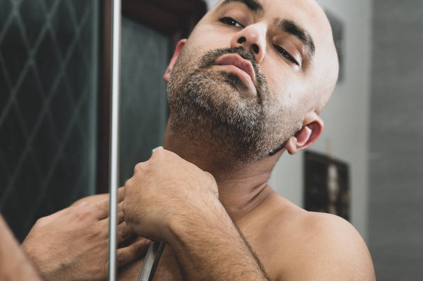Un hombre asiático británico calvo, arreglando y esculpiendo su barba en casa en el baño, frente al espejo. Está usando una navaja de afeitar tradicional o una navaja de barbero. Esto se puede utilizar para temas relacionados con la masculinidad, citas y la mediana edad
.  - Foto, imagen