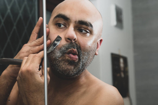 A bald, British Asian man, grooming and sculpting his beard at home in the bathroom, in front of the mirror. He is using a traditional cut throat razor or a barber's razor. This can be used to theme issues around maleness, dating and middle age.  - Fotoğraf, Görsel