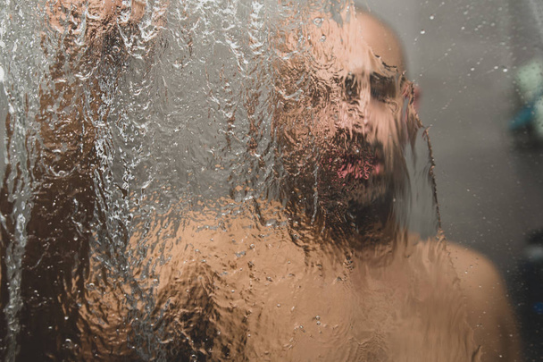 British Asian man taking a shower in the shower cubicle - with copy space. Bald middle aged man in shower cubicle, taking a shower. The facial expressions can be used to convey feelings such as sadness, worry, depression and loneliness.  - Foto, afbeelding