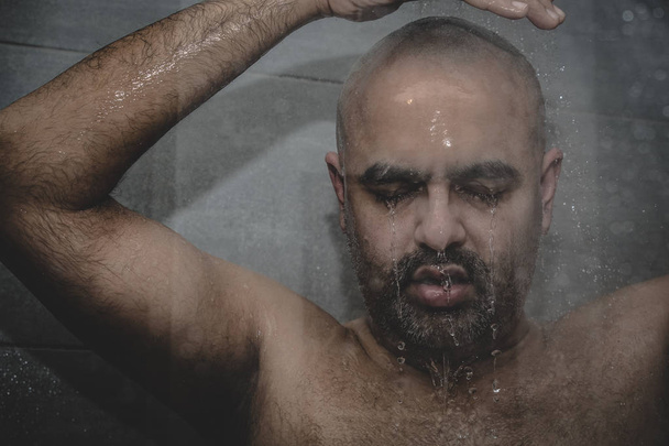 British Asian man taking a shower in the shower cubicle - with copy space. Bald middle aged man in shower cubicle, taking a shower. The facial expressions can be used to convey feelings such as sadness, worry, depression and loneliness.  - Photo, Image
