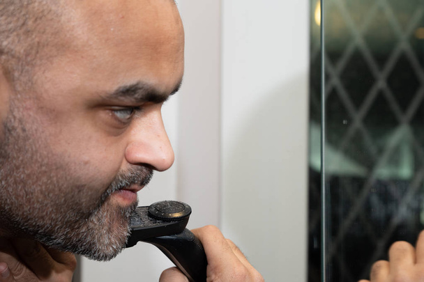 イギリスのアジア人男性、無精ひげ、鏡の前にひげをトリミング. - 写真・画像