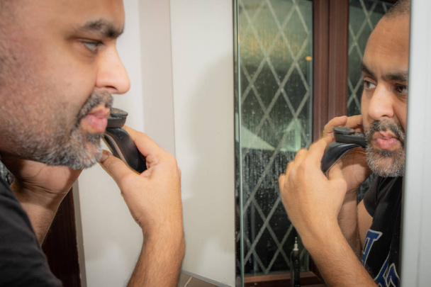 イギリスのアジア人男性、無精ひげ、鏡の前にひげをトリミング. - 写真・画像