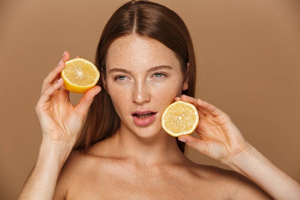 Beauty image of european shirtless woman with long hair holding two pieces of orange isolated over beige background - Φωτογραφία, εικόνα