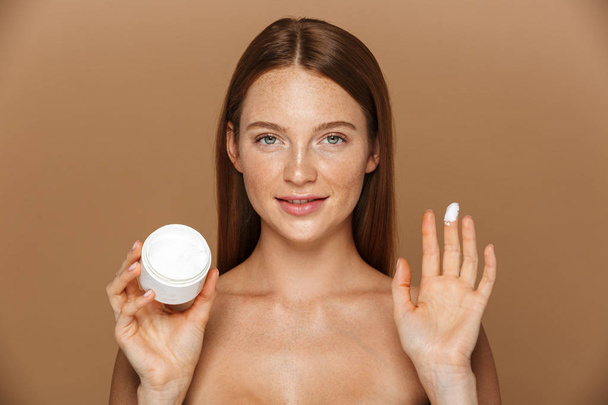Beauty image of brunette shirtless woman smiling and holding jar with face cream isolated over beige background - Foto, afbeelding
