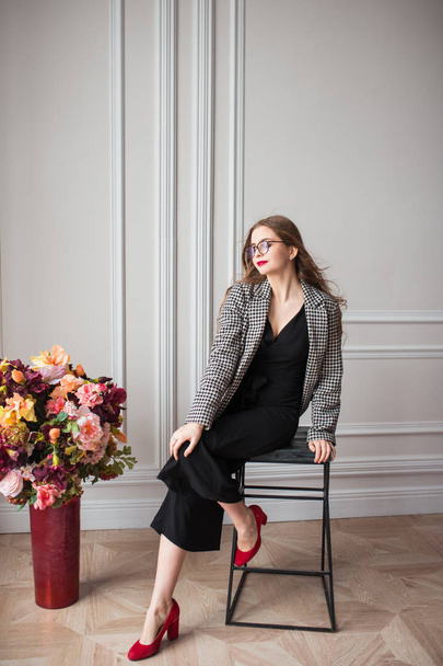 fiatal gyönyörű divat modell fekete ruha, elegáns blézer és szemüveg ül a széken, beltéri virágok - Fotó, kép