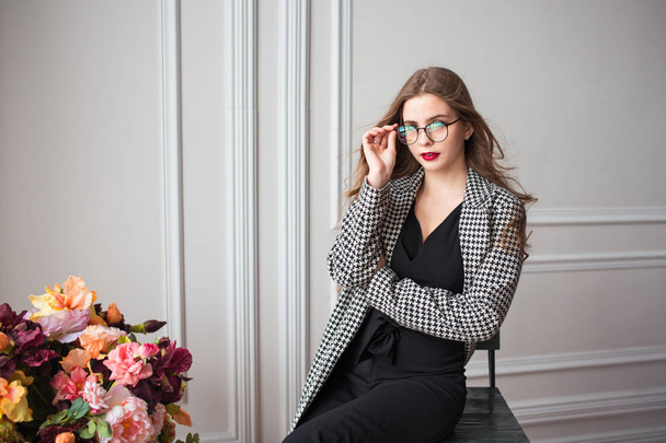 modelo de moda hermosa joven en vestido negro y blazer elegante y gafas sentado en silla con flores en el interior
 - Foto, Imagen