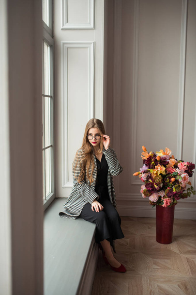 fiatal, érzéki nő fekete ruha, elegáns blézer és szemüveg pózol a ablakpárkányon beltéri - Fotó, kép