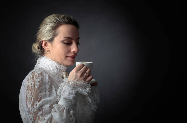 Retrato de una mujer vestida de victoriano con una taza de café. Blusa blanca con encaje, bordado y cuello alto. Fondo negro. Copiar espacio
. - Foto, imagen