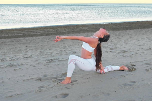 Young woman is doing yoga High Lunch Pose on the sand beach at sunrise in autumn. - Foto, afbeelding