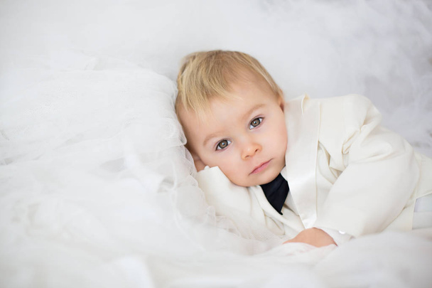 Portrait of elegant handsome little boy in a tuxedo, lying in bed on his moms bridal dress, ready to go to wedding - Fotoğraf, Görsel