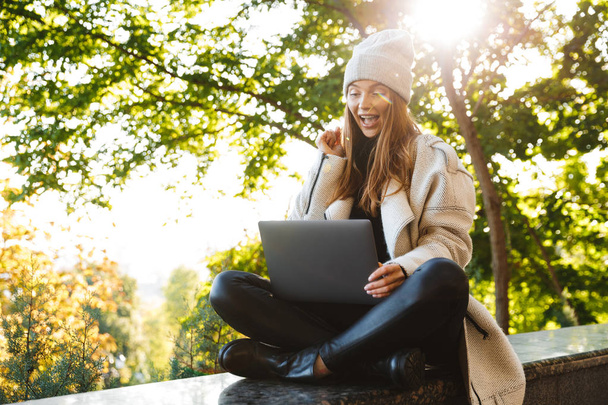 Beautiful young woman dressed in autumn coat and hat sitting outdoors, using laptop computer, celebrating success - Foto, Imagen