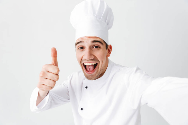 Happy chef cook wearing uniform standing isolated over white background, taking a selfie, giving thumbs up - 写真・画像