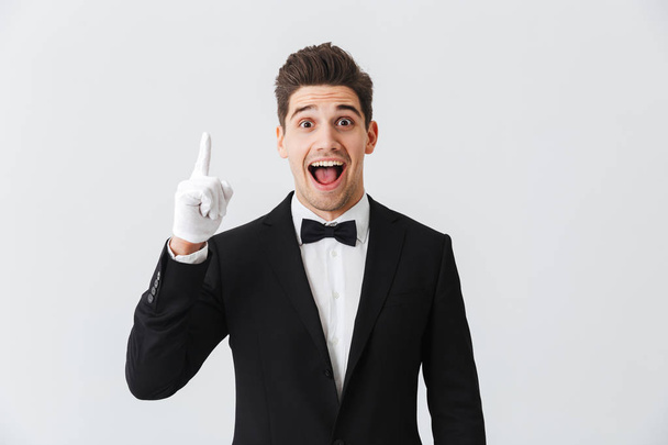 Portrait of a handsome young man waiter wearing tuxedo and gloves standing isolated over white background, pointing up - Photo, Image