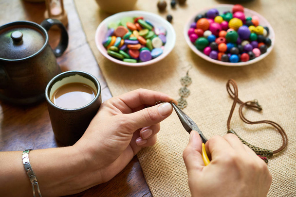 Gros plan de la femme assise à la table buvant du café et fabriquant des accessoires faits à la main à partir de perles colorées
 - Photo, image