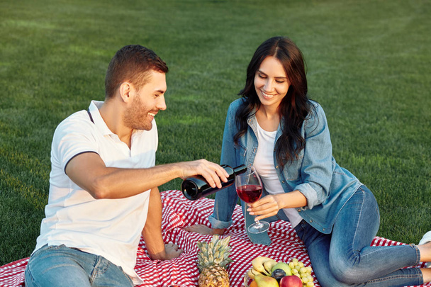 man pouring red wine from the bottle in the glass for girlfriend at picnic in the park - Foto, Imagem