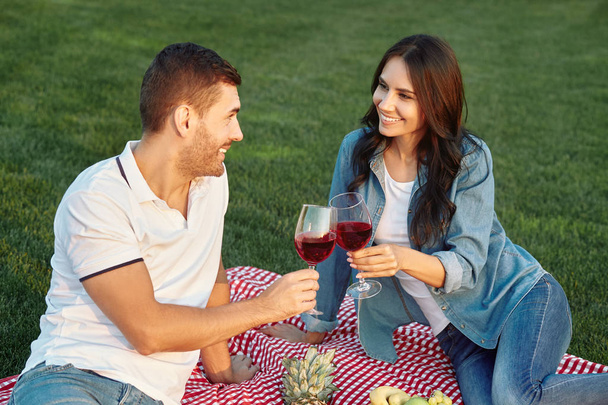 couple in love loojking at each other, having small picnic with fruits and clicking with wine glasses sitting on the blanket in the park - Fotó, kép