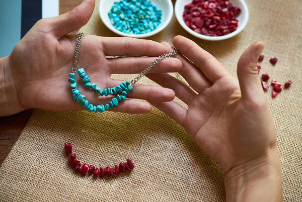 Primer plano de las manos femeninas sosteniendo su collar trabajado a mano de piedras naturales, y tejiendo el otro en la mesa
 - Foto, imagen