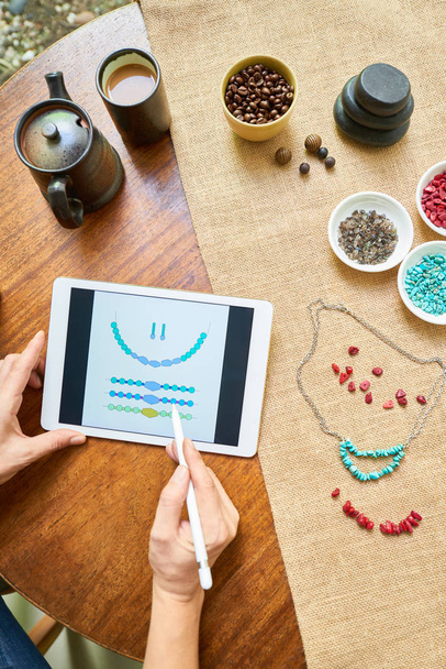 Frau, die am Tisch sitzt und die Perlen-Handy-App nutzt, fertigt sie ihre Halskette in Handarbeit nach dem Muster - Foto, Bild