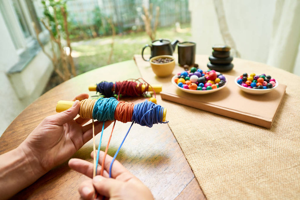 Primer plano de la mujer sentada en la mesa y haciendo pulseras de cuerdas y cuentas de colores
 - Foto, Imagen
