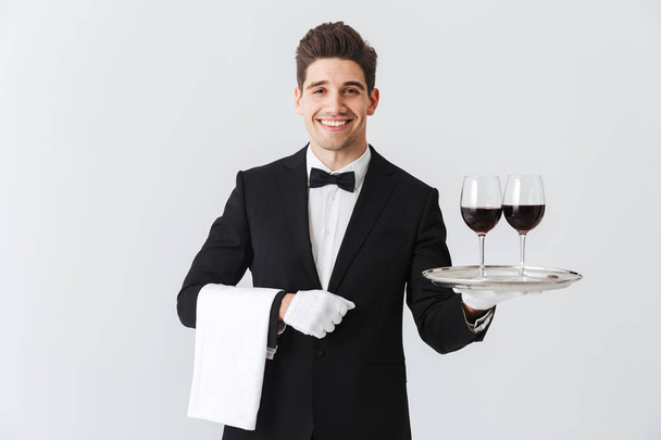 Handsome young waiter wearing tuxedo presenting a tray with two glasses of red wine isolated over gray background - Photo, image