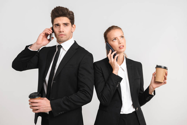Confident young business couple standing isolated over gray background, holding cups of takeaway coffee, using mobile phones, talking - Foto, immagini
