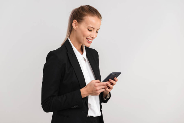 Sorridente donna d'affari bionda in piedi isolato su sfondo grigio, utilizzando il telefono cellulare
 - Foto, immagini